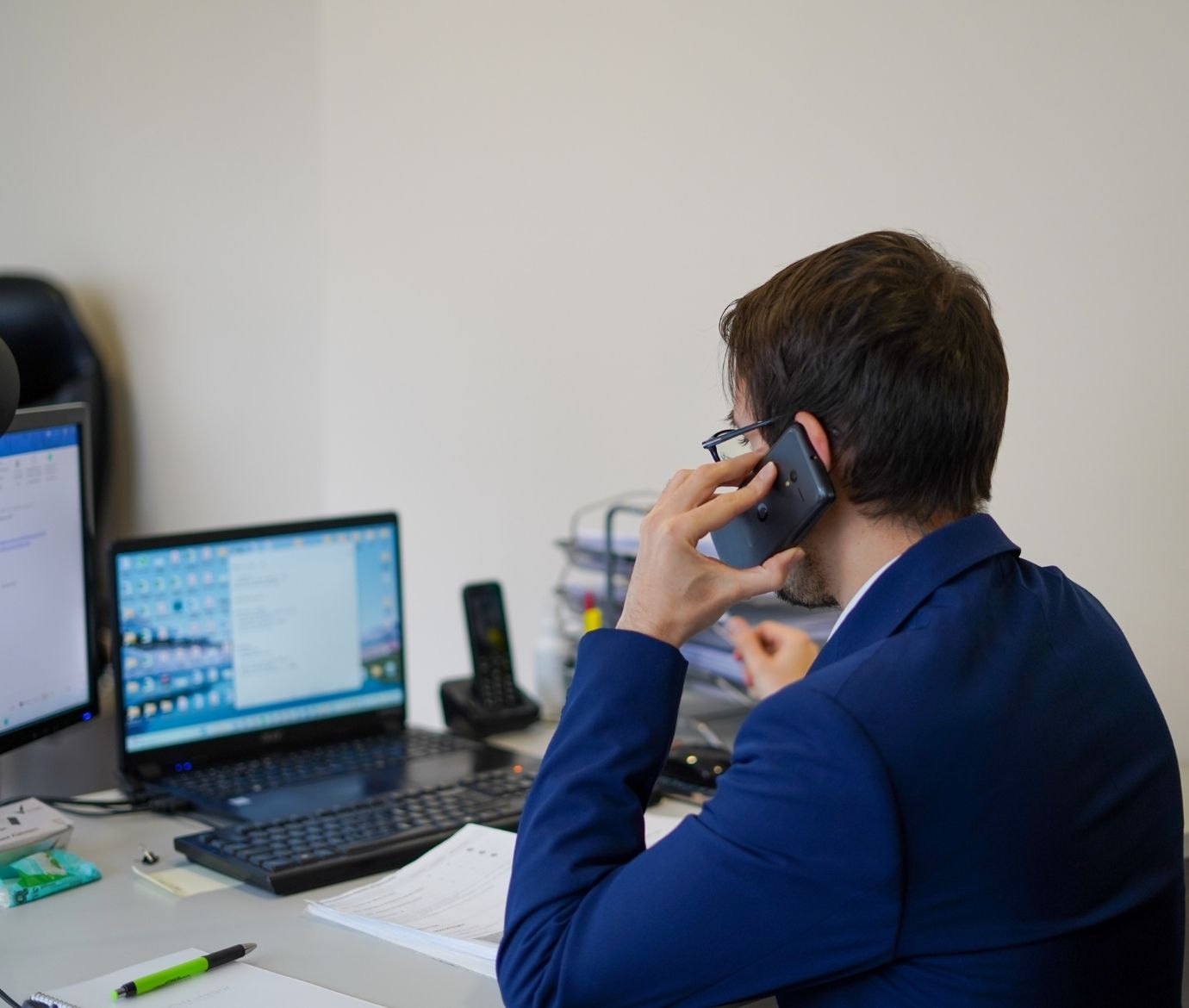 Vista lateral traseira de um homem com óculos e fato azul, sentado numa secretária de escritório enquanto fala ao telemóvel. À sua frente, encontram-se um computador portátil aberto, um monitor adicional, um teclado e vários documentos organizados.